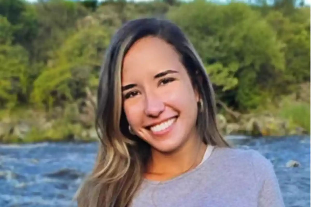 A person smiling outdoors with a river and trees in the background.