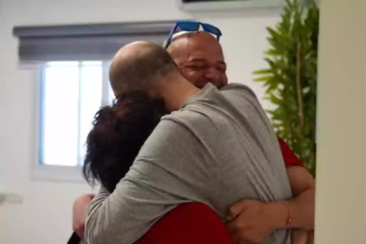 Three people embrace joyfully in a room lit by a window.