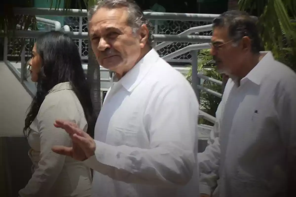 A gray-haired man with a mustache, wearing a white jersey, walks alongside two other people in an outdoor setting.