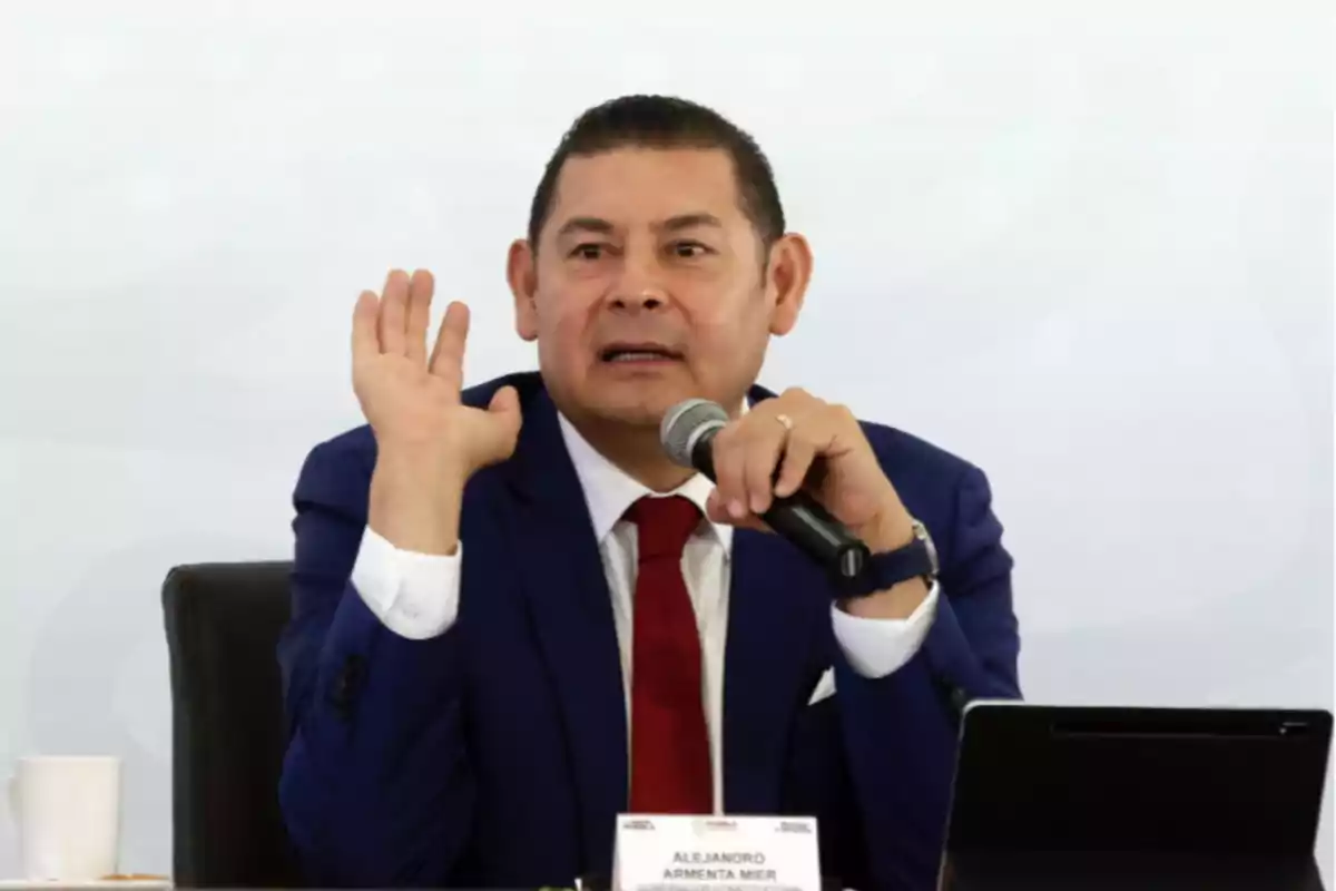 A man in a blue suit and red tie speaks into a microphone while raising one hand.