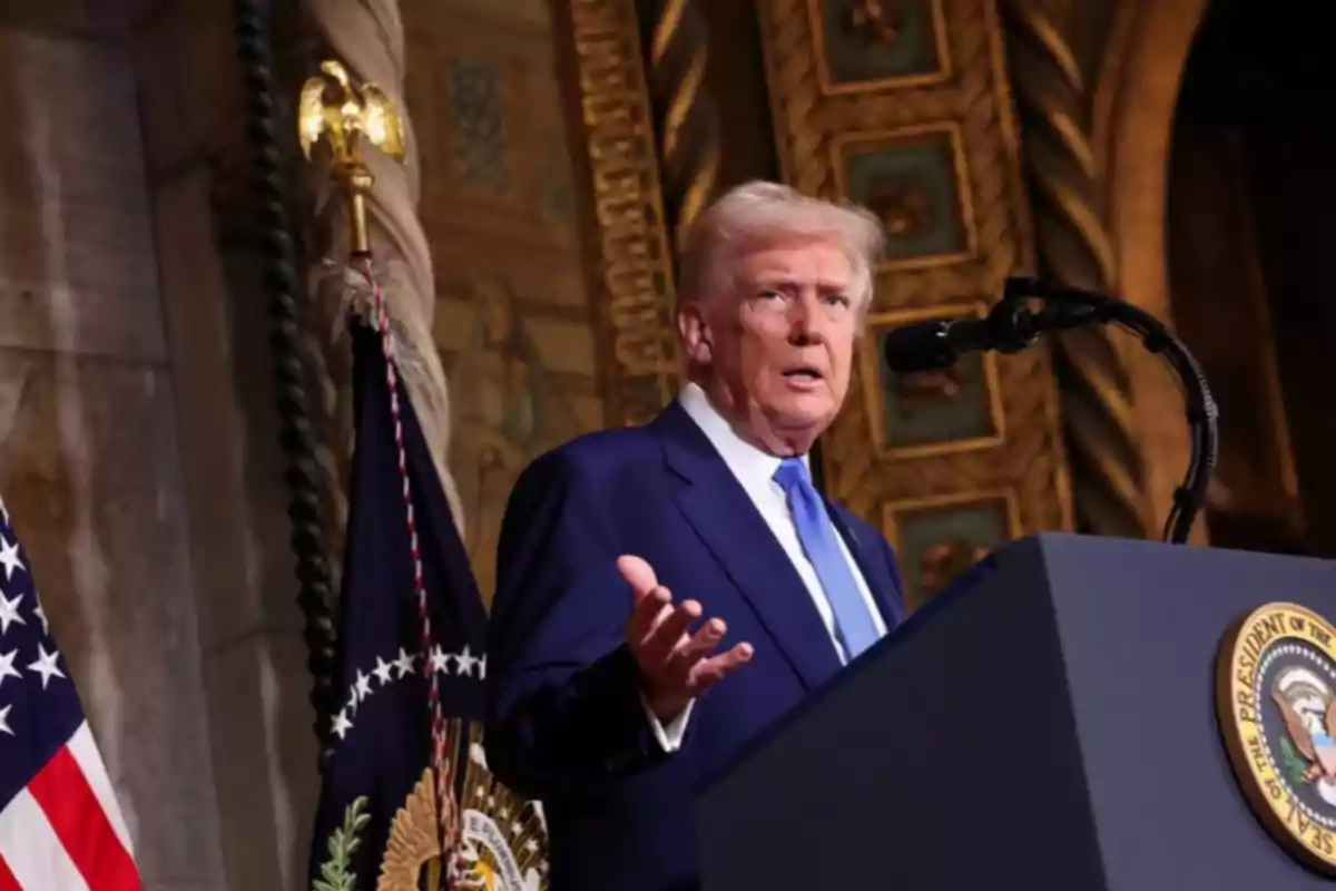 A man in a blue suit speaks in front of a podium with a microphone, with flags and a decorative background behind him.