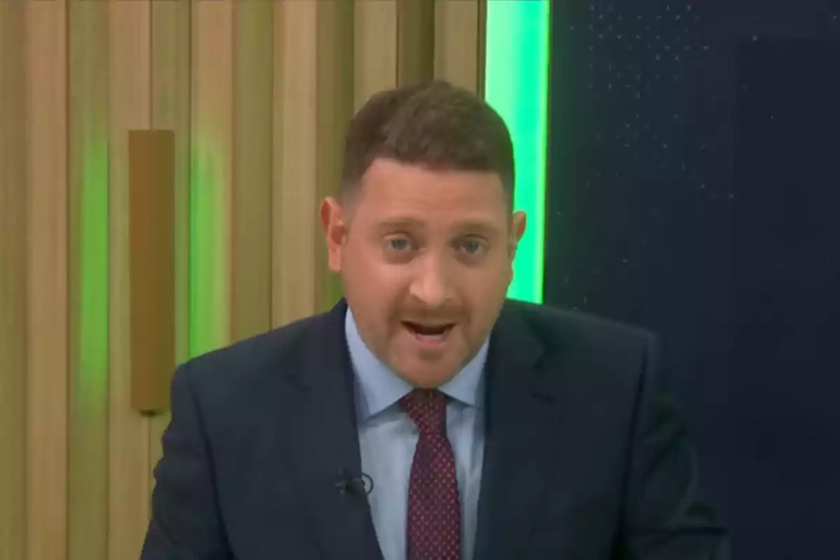 A man in a suit and tie speaks in front of a wood-paneled wall and a green light.