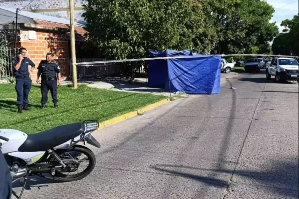 A street scene with police officers, a motorcycle, and a blue tent in a cordoned-off area.