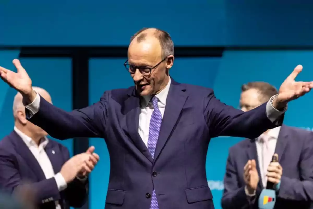 A man in a suit and blue tie raises his arms while other people applaud in the background.