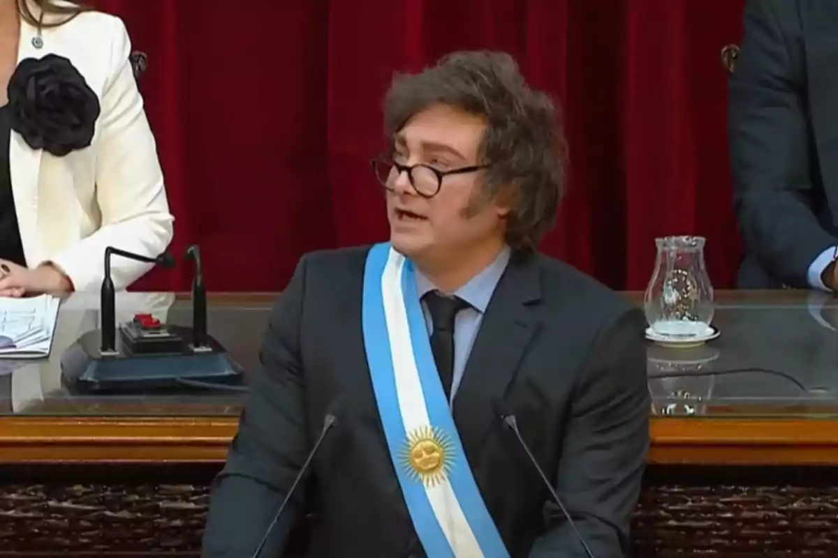 Un hombre con gafas y una banda presidencial habla en un podio frente a un fondo rojo.