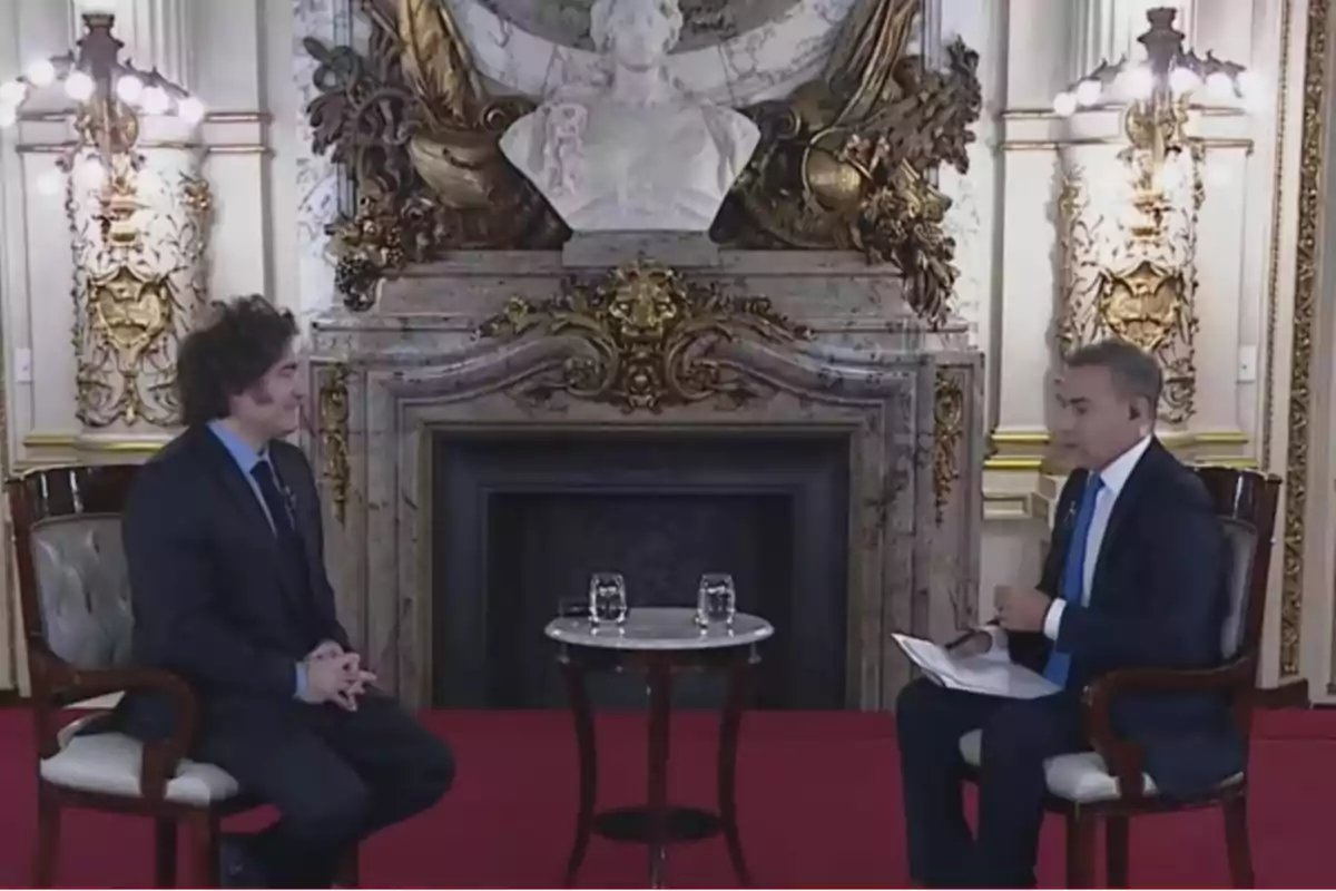 Dos personas sentadas en una elegante sala con decoración clásica y una chimenea de fondo, conversan frente a una mesa pequeña con vasos de agua.