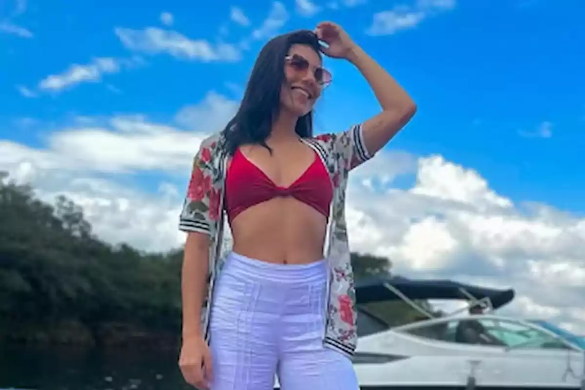 Mujer sonriendo con gafas de sol y ropa veraniega, posando frente a un bote y un cielo azul con nubes.