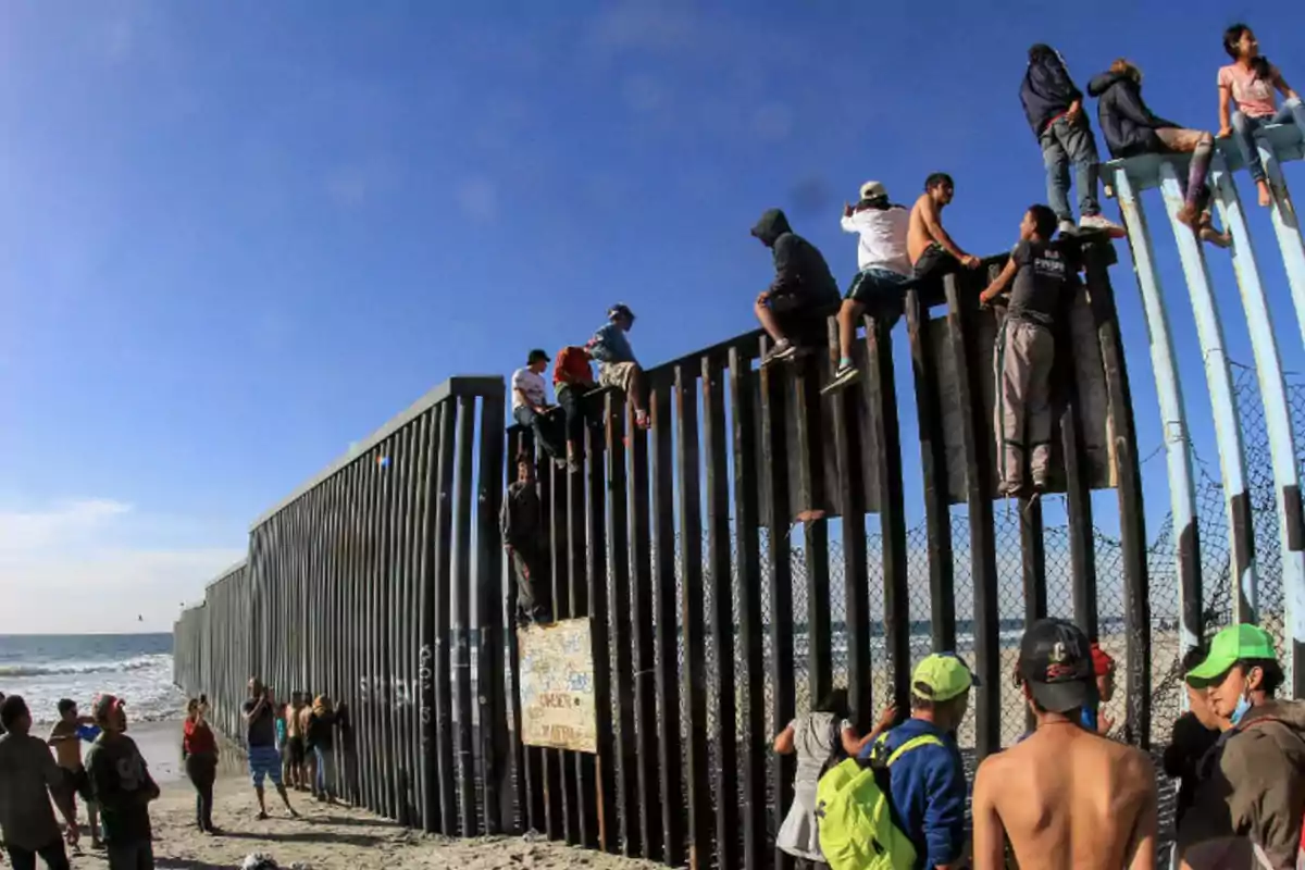 People climbing a metal fence on a beach under a clear sky.