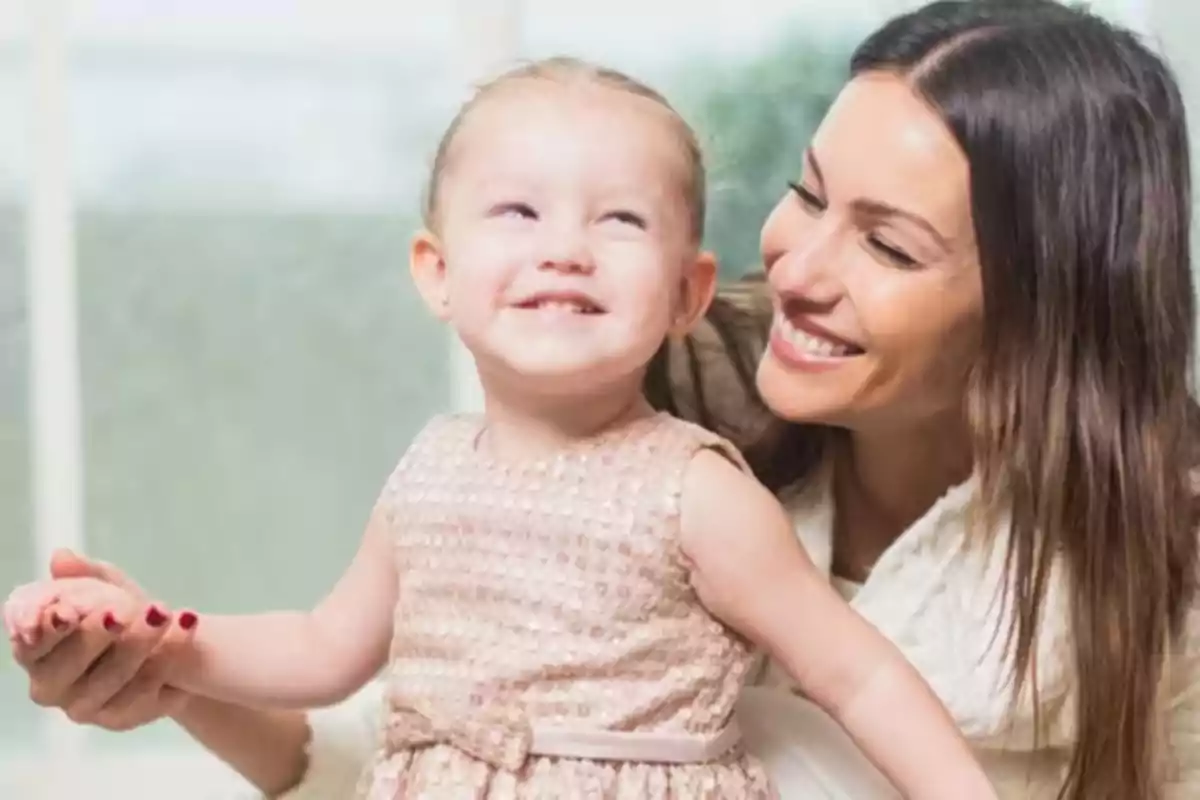 Una mujer sonriente sostiene la mano de una niña pequeña que también sonríe.