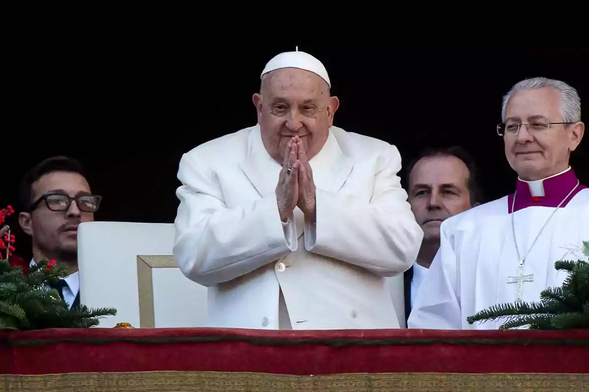 El papa Francisco vestido de blanco con las manos juntas en un gesto de oración acompañado de otras personas en un balcón decorado con plantas.