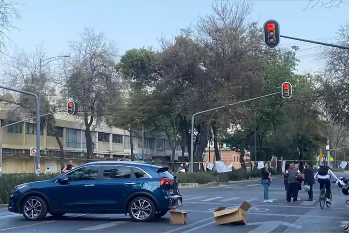 Un coche azul detenido en un cruce con semáforos en rojo y un grupo de personas y un ciclista cerca, con cajas de cartón en el suelo.