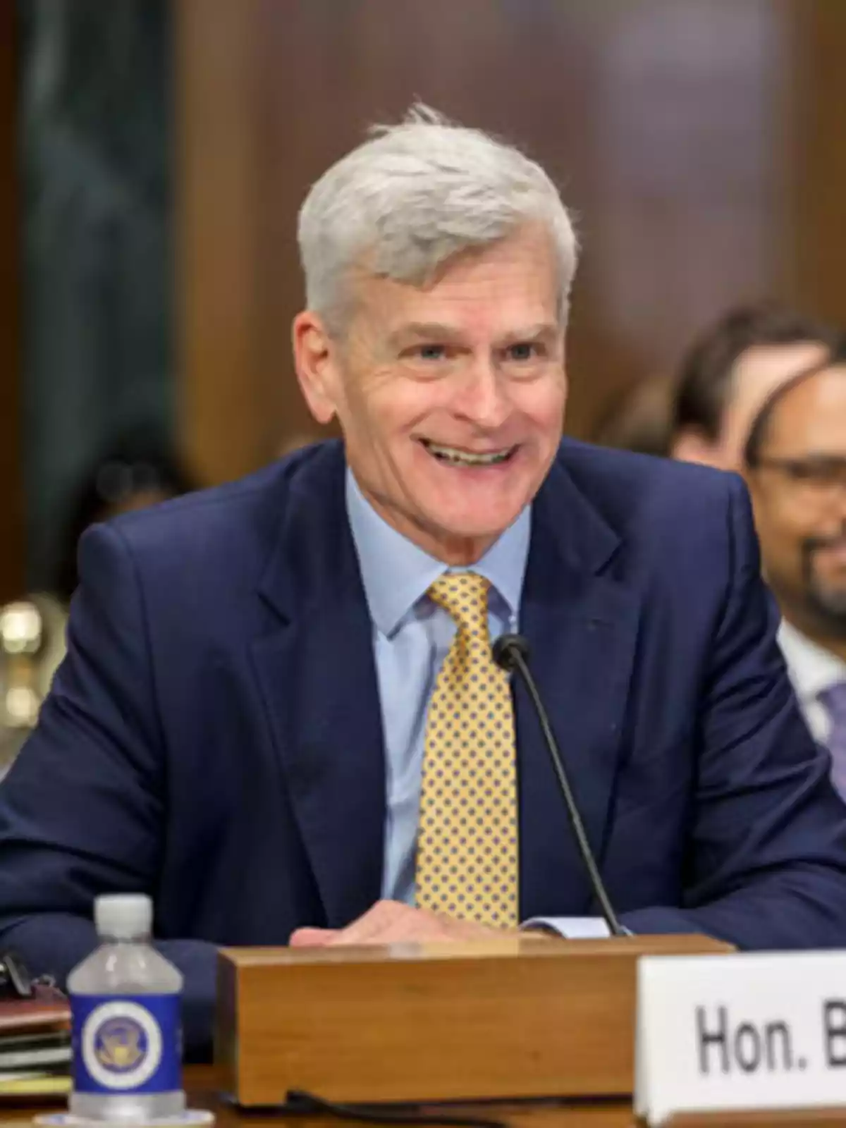 A gray-haired man in a blue suit smiles while sitting at a table with a microphone in front of him.