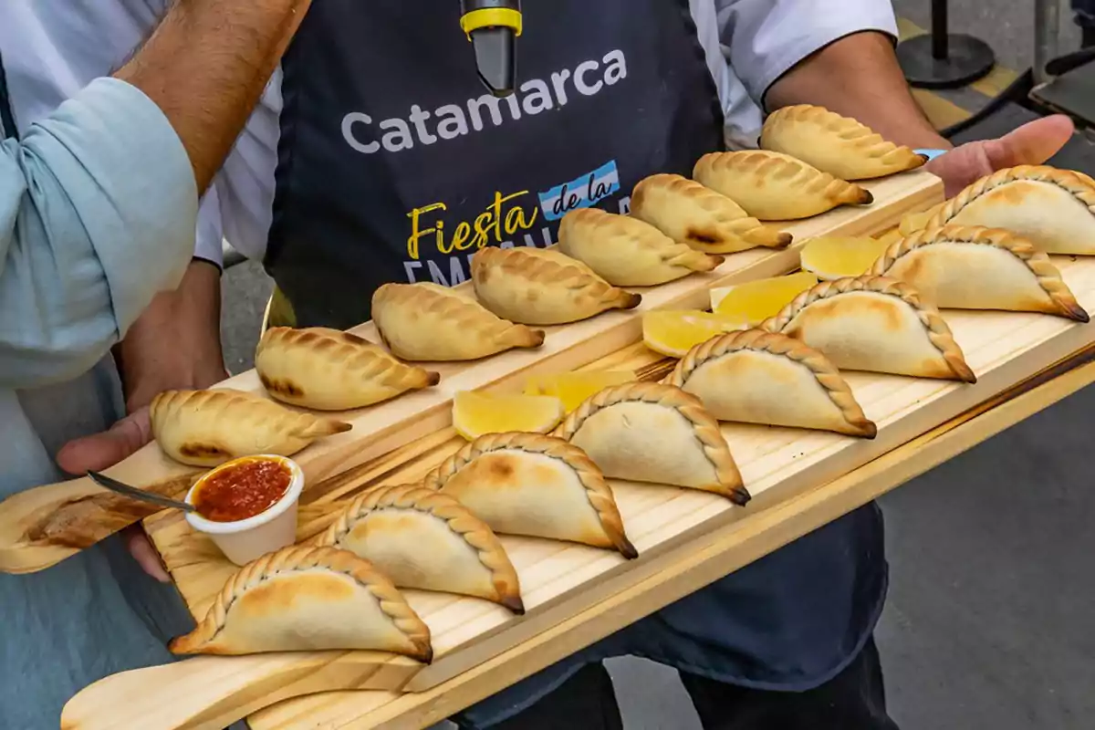 Una persona sostiene una bandeja de madera con varias empanadas horneadas acompañadas de rodajas de limón y un recipiente pequeño con salsa roja, mientras viste un delantal que dice "Catamarca Fiesta de la Empanada".