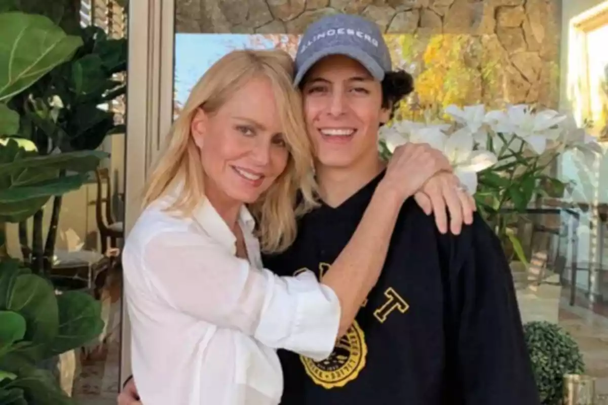 A blonde woman hugs a young man wearing a cap and a black sweatjersey, both smiling in front of a house with plants and white flowers.