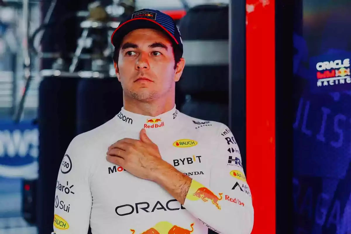 Un piloto de carreras con uniforme blanco y gorra está de pie con la mano en el pecho en un entorno de boxes de automovilismo.