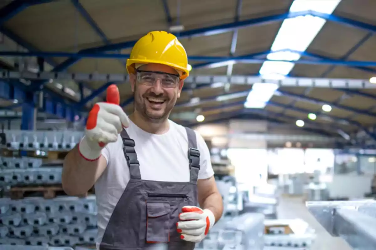 Trabajador en una fábrica con casco amarillo y gafas de seguridad sonriendo y levantando el pulgar.