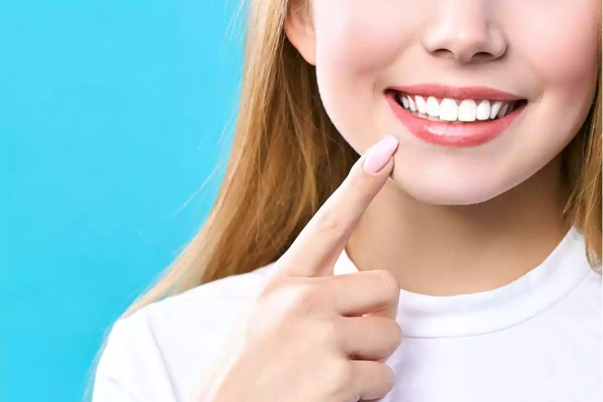 Una persona sonriendo y señalando sus dientes blancos sobre un fondo azul.