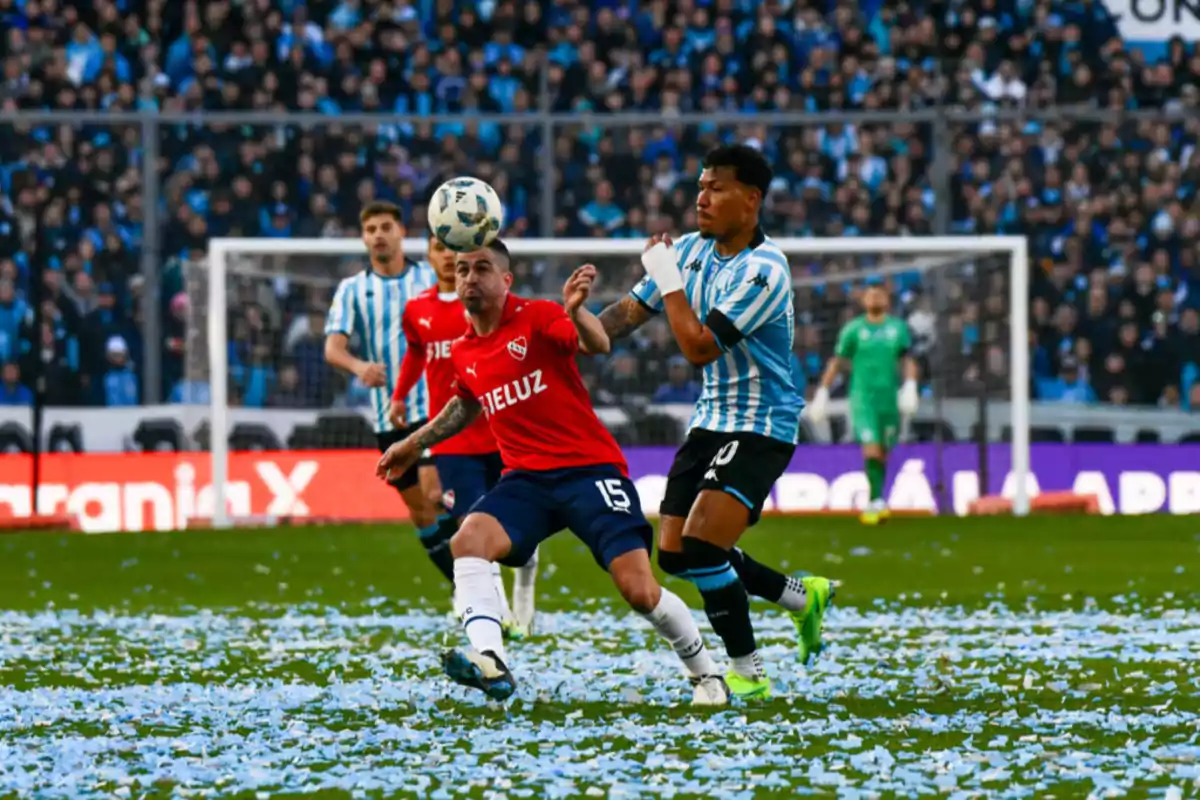 Jugadores de fútbol compiten por el balón en un partido, con un estadio lleno de espectadores y confeti en el campo.