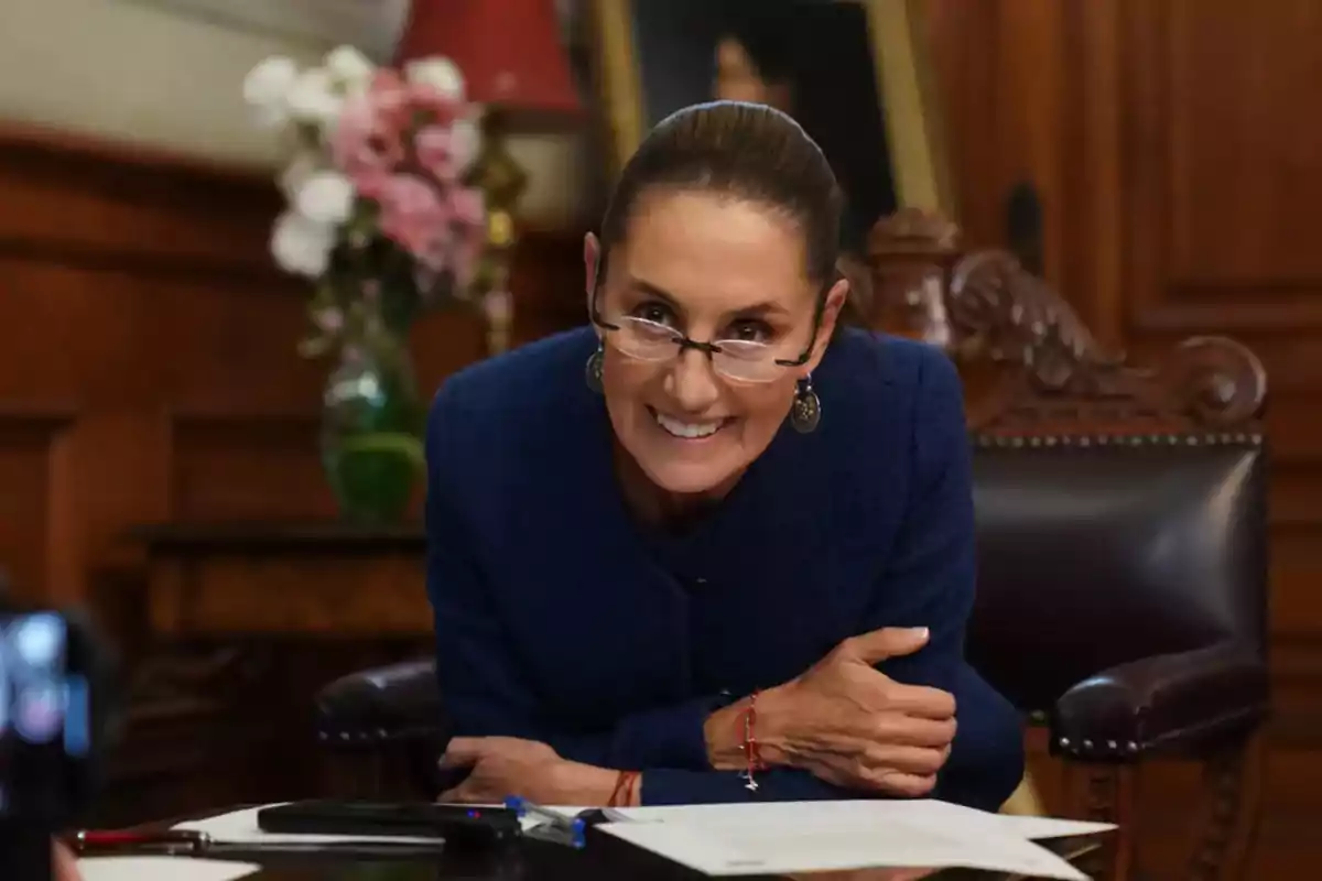 Una mujer sonriente con gafas y cabello recogido está sentada en una oficina elegante con flores en el fondo.