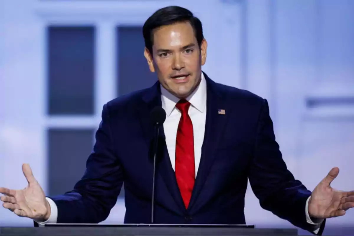 A man in a dark suit with a red tie speaks at a podium with a microphone against a light blue background.