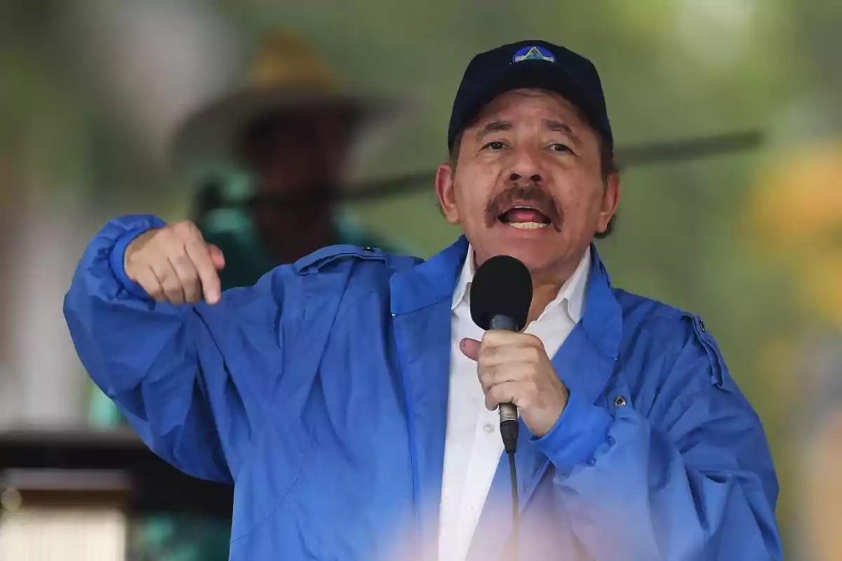 Un hombre con chaqueta azul y gorra habla en un micrófono durante un evento al aire libre.