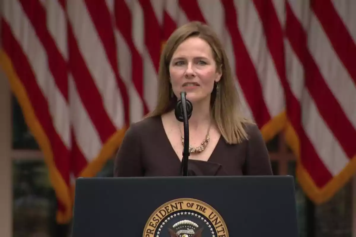 Una mujer hablando en un podio con el sello presidencial de Estados Unidos y banderas estadounidenses de fondo.