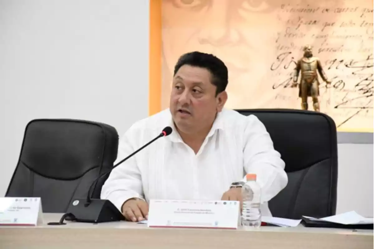 A man in a white jersey speaks into a microphone during a meeting, with a bottle of water and documents on the table.