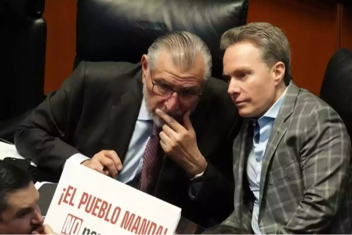 Dos hombres en traje conversan en un entorno formal, uno de ellos sostiene un cartel que dice "¡El pueblo manda!"