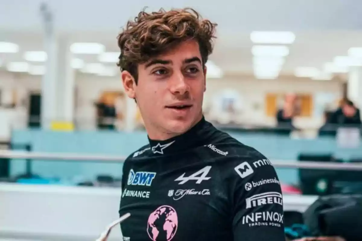 Un hombre con cabello rizado lleva un traje de carreras negro con varios logotipos en un entorno interior.