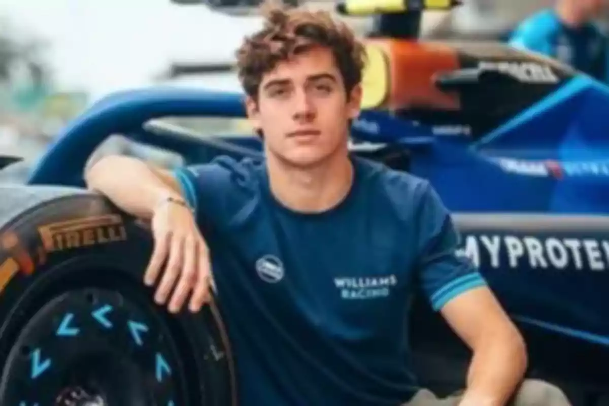 Joven con camiseta de Williams Racing sentado junto a un auto de carreras.