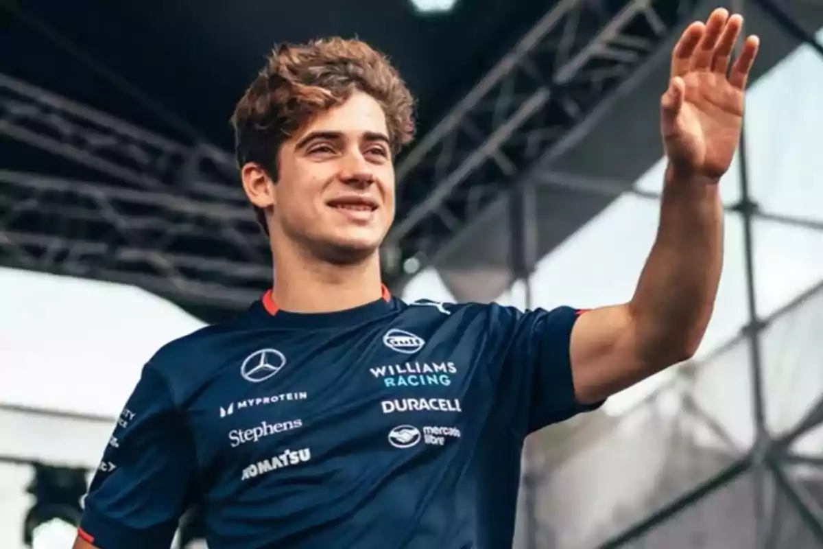 Un hombre joven con camiseta de Williams Racing saluda con la mano en un escenario al aire libre.