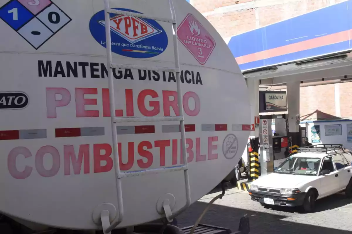 Un camión cisterna con advertencias de peligro de combustible inflamable está estacionado en una estación de gasolina donde se observa un automóvil blanco.