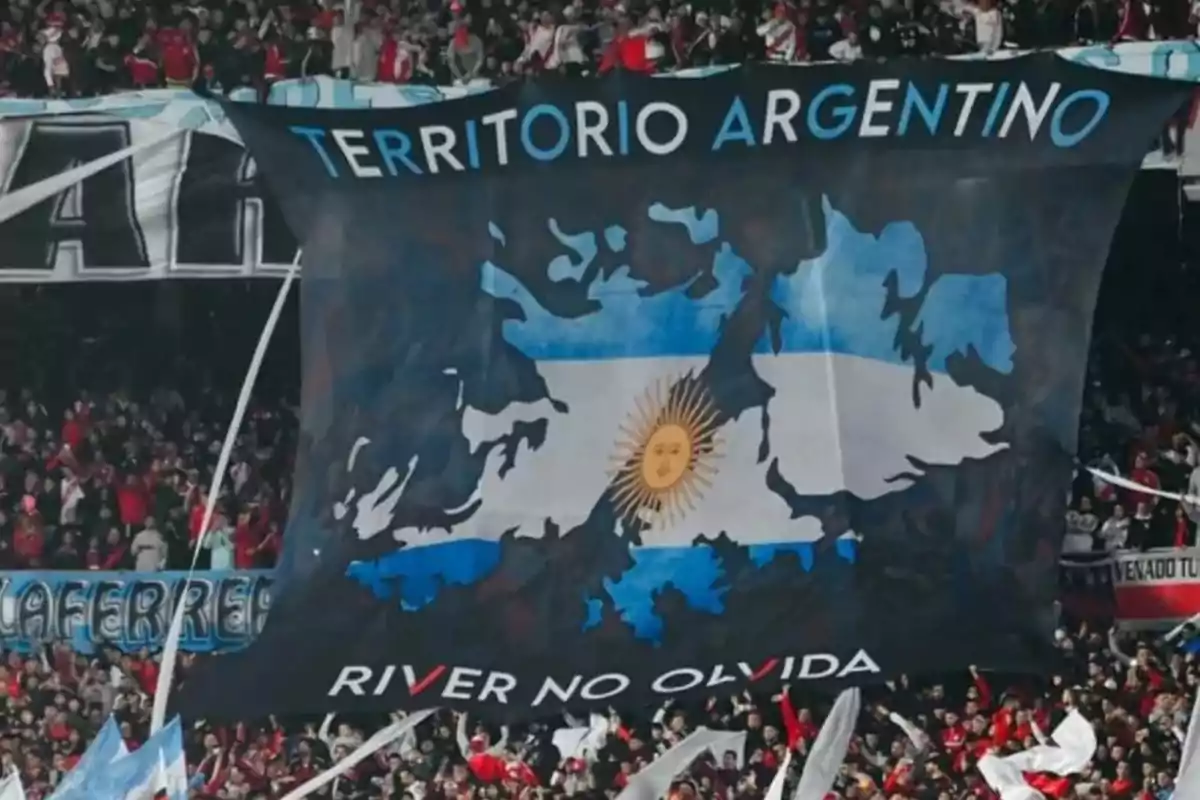 Una bandera con el mapa de las Islas Malvinas y los colores de la bandera argentina, con las palabras "Territorio Argentino" en la parte superior y "River no olvida" en la parte inferior, sostenida por una multitud de personas en un estadio.