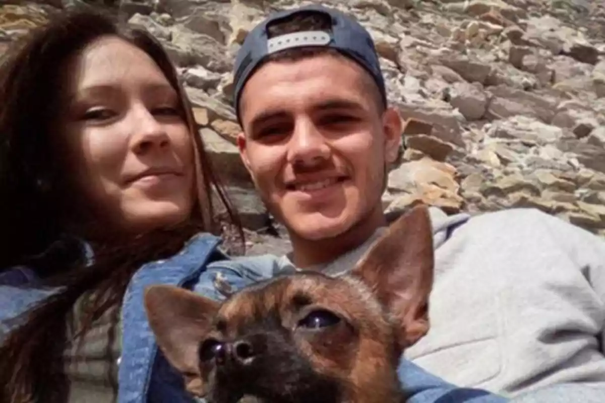 Una pareja joven sonríe mientras sostiene a un pequeño perro frente a un fondo de piedras.