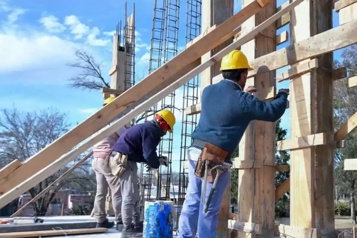 Trabajadores de la construcción con cascos amarillos trabajando en una estructura de madera y metal bajo un cielo despejado.