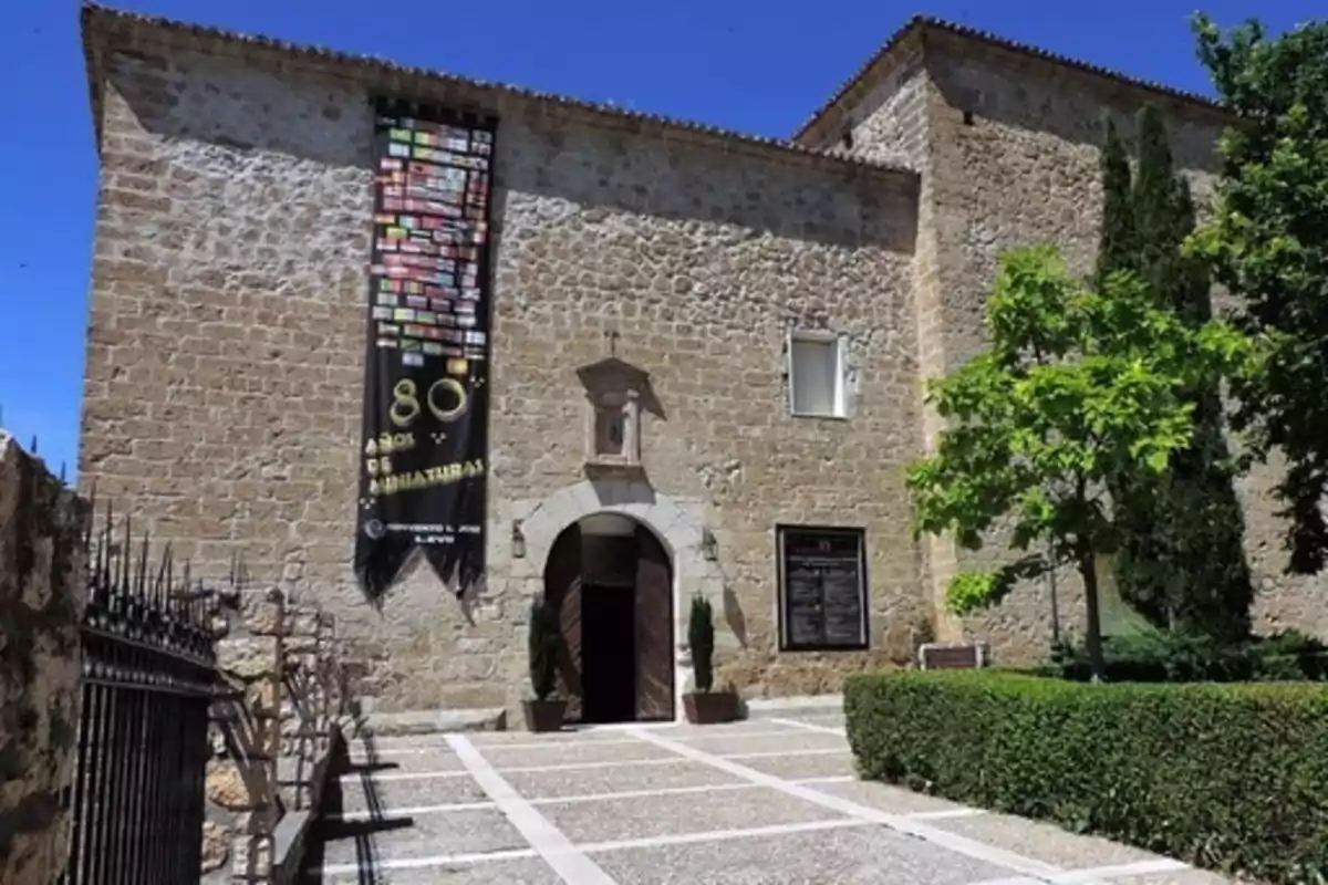 Edificio de piedra con una gran pancarta negra colgada en la fachada, rodeado de árboles y arbustos, con una entrada arqueada y un camino pavimentado al frente.