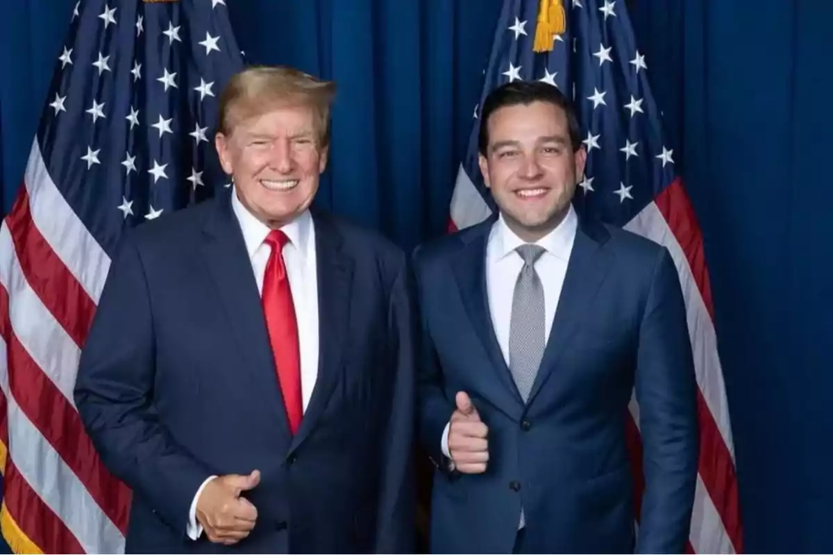 Dos hombres de traje posan sonriendo frente a banderas estadounidenses.