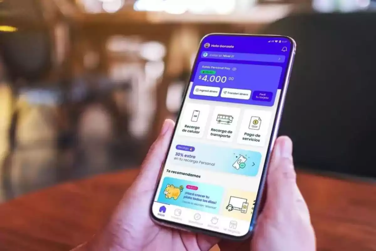 Persona sosteniendo un teléfono móvil que muestra una aplicación de gestión financiera con opciones de recarga de celular, transporte y pago de servicios.