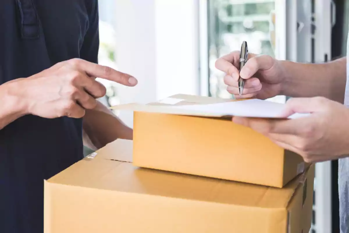 Dos personas interactúan durante la entrega de un paquete, una firma un documento sobre una caja de cartón mientras la otra señala.