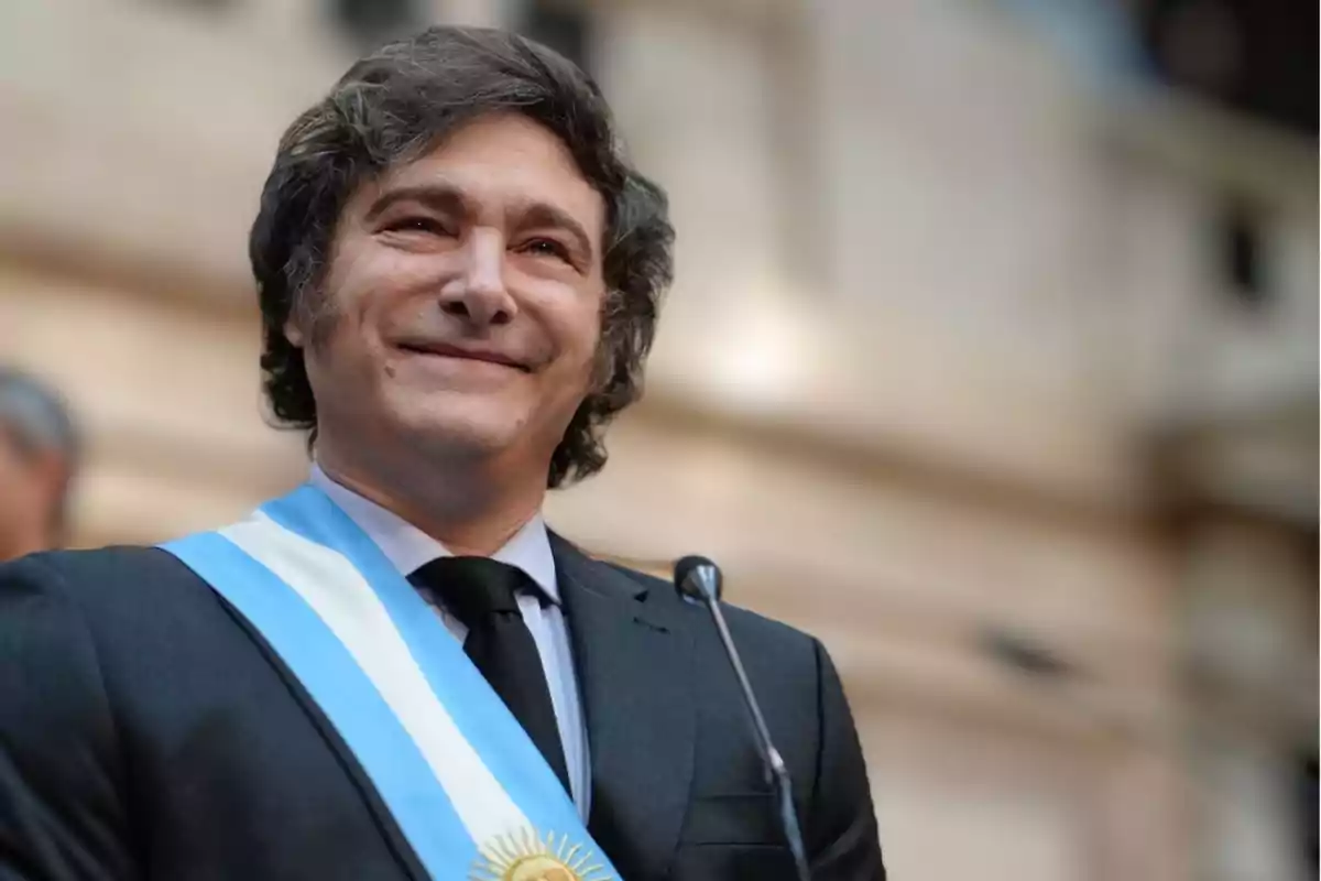 Un hombre con traje oscuro y corbata negra lleva una banda presidencial azul y blanca mientras sonríe frente a un micrófono.