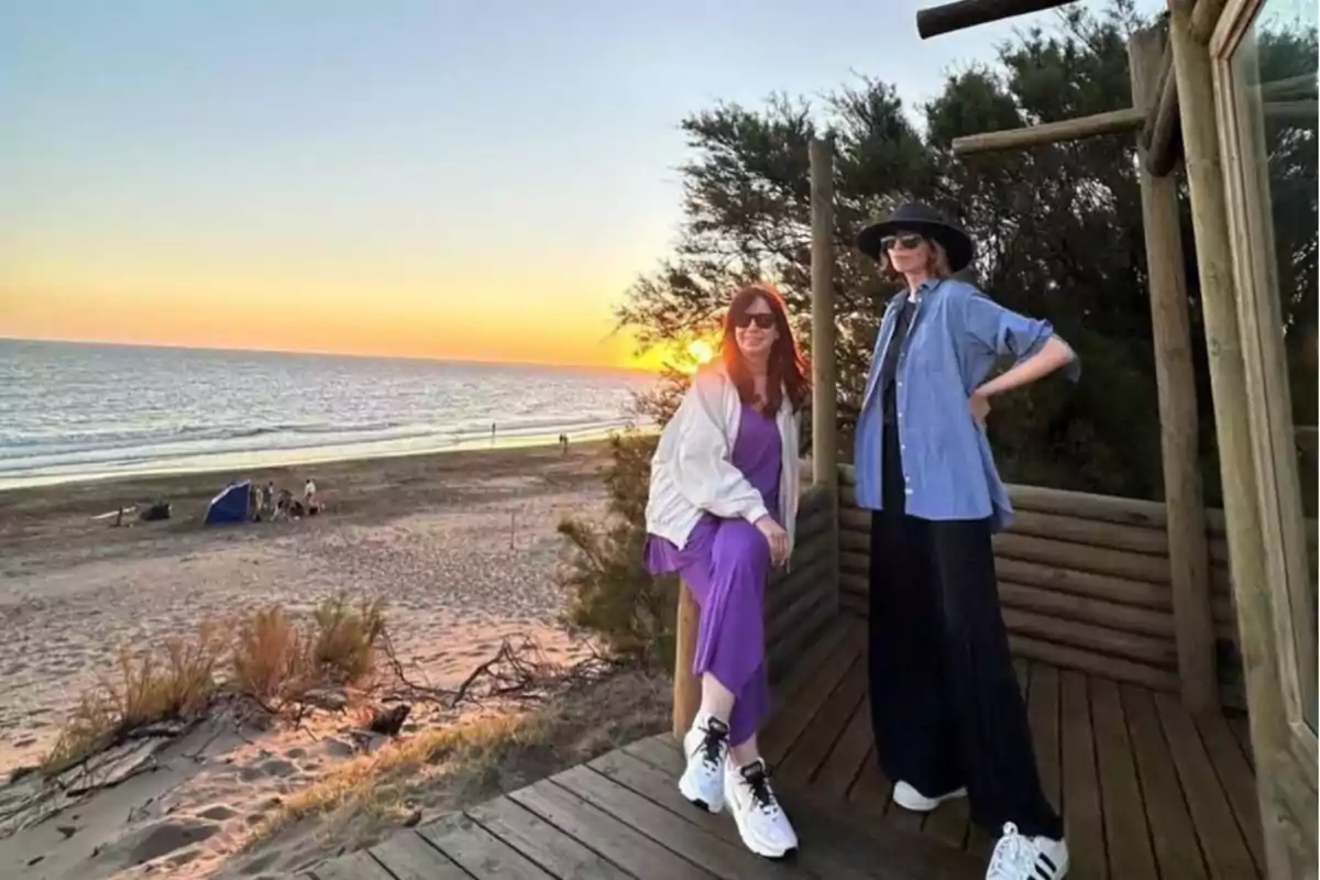 Dos personas posan en una plataforma de madera junto a la playa al atardecer con el mar y el cielo colorido de fondo.