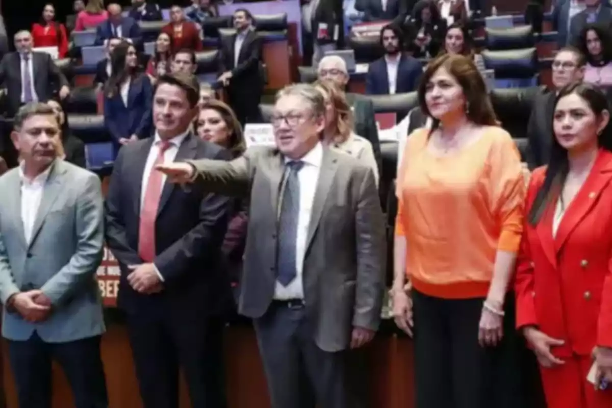 Un grupo de personas de pie en un auditorio, algunas de ellas vestidas formalmente, con un hombre en el centro señalando hacia adelante.