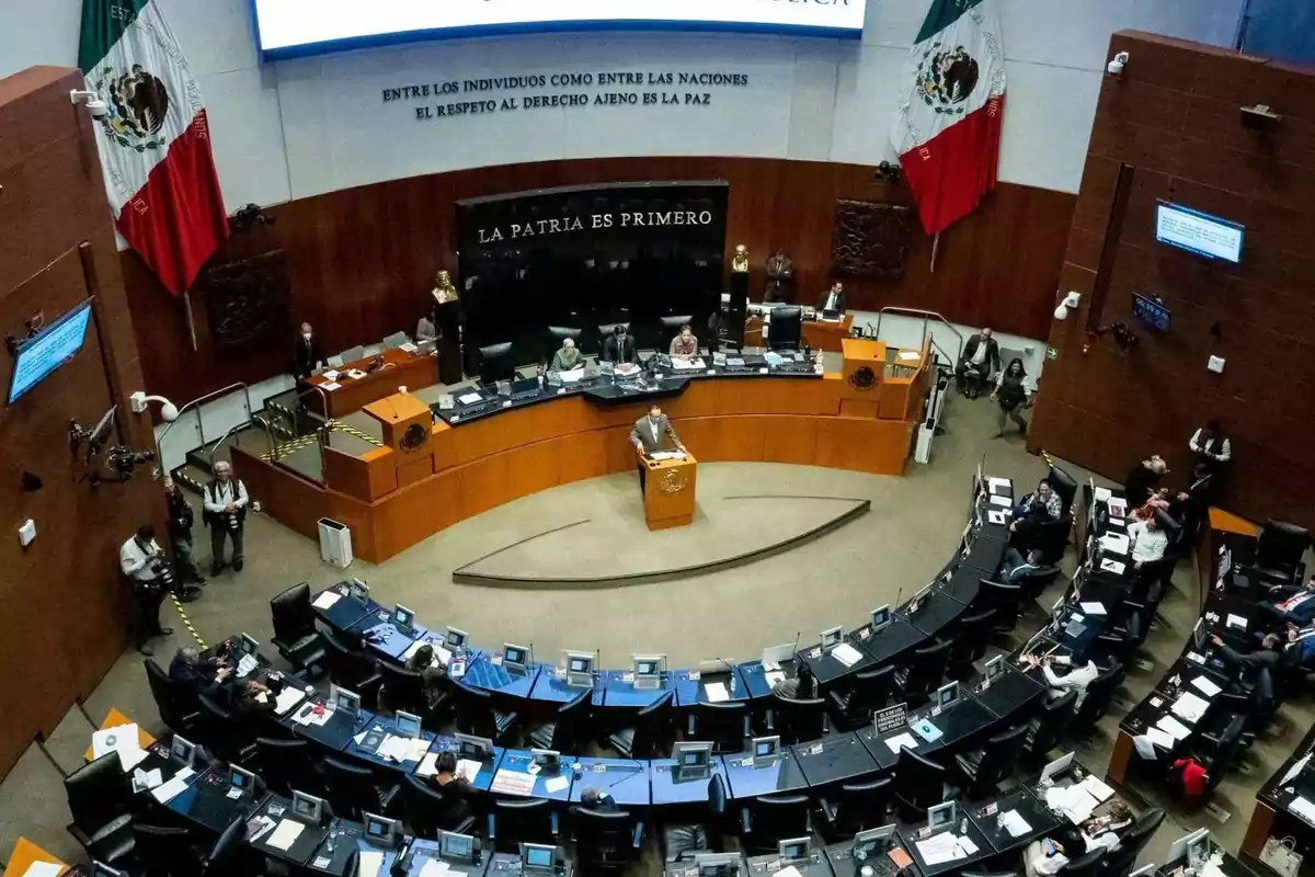 Una vista aérea del interior de un recinto legislativo con personas sentadas en escritorios semicirculares y una persona hablando en un podio, con banderas y un lema en la pared.