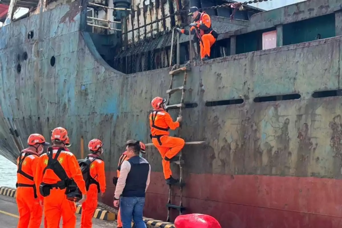 Personas con uniformes naranjas suben por una escalera de cuerda a un barco oxidado.