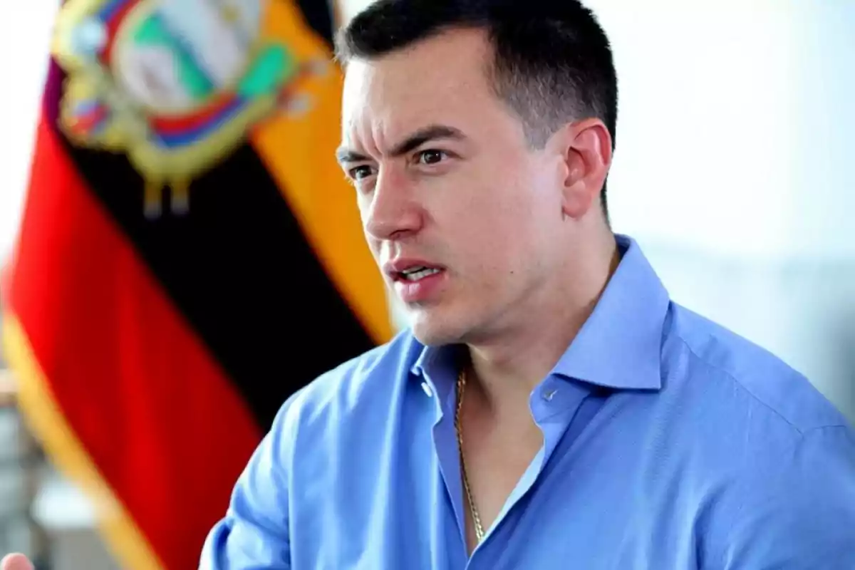 Un hombre con camisa azul hablando frente a una bandera de Ecuador.