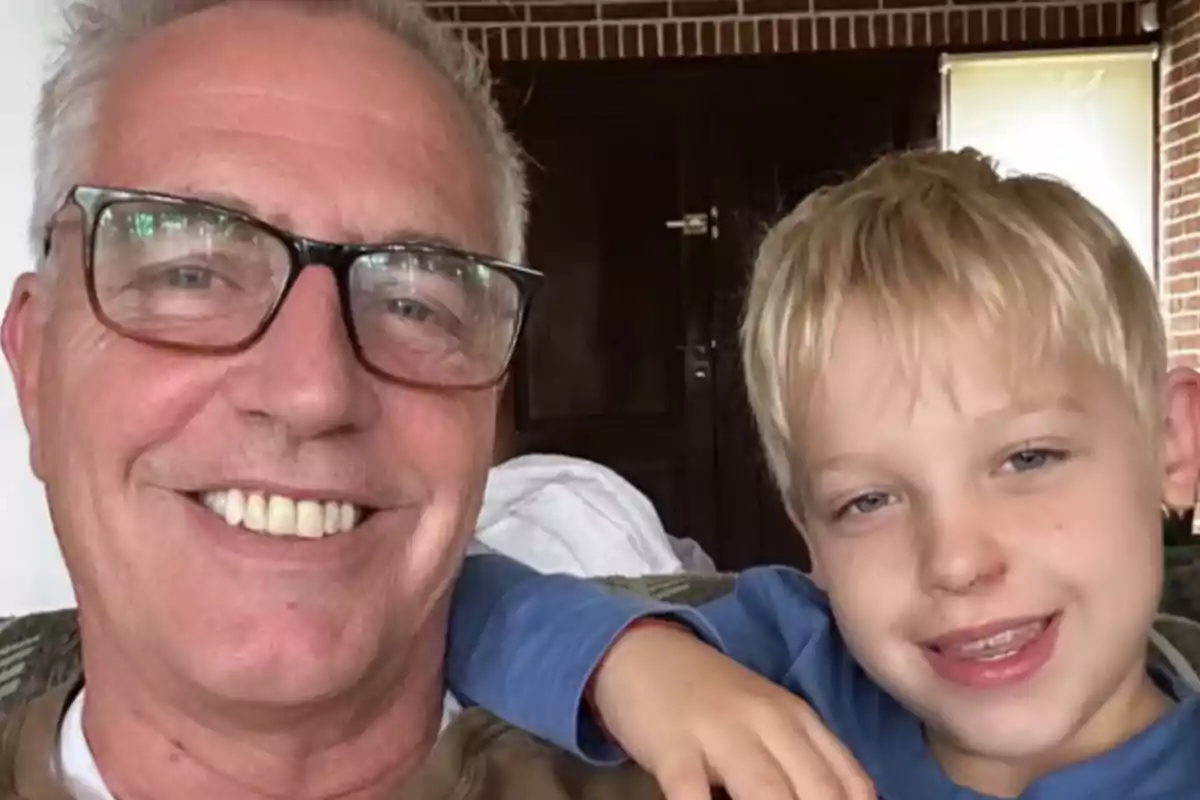 An older man with glasses and a blond child smile together indoors.