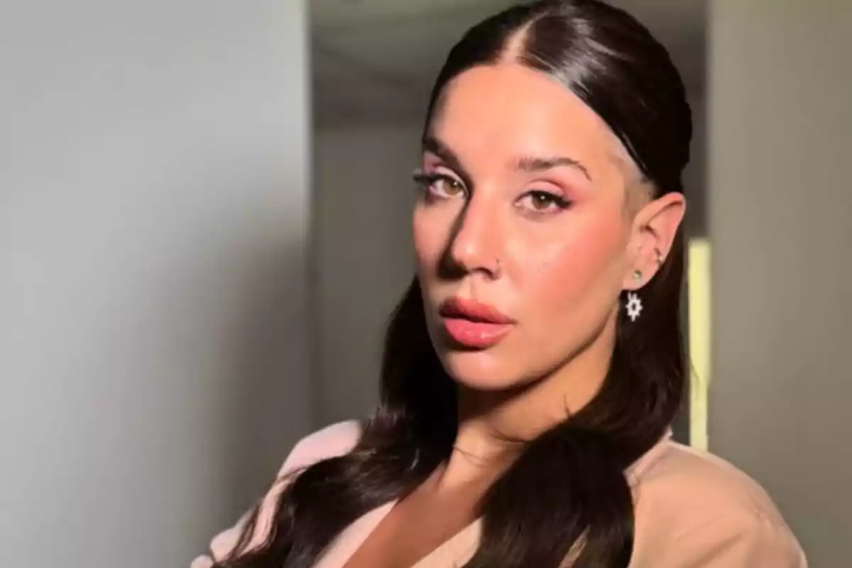 A person with long dark hair, wearing earrings and subtle makeup, looks at the camera in an indoor setting.