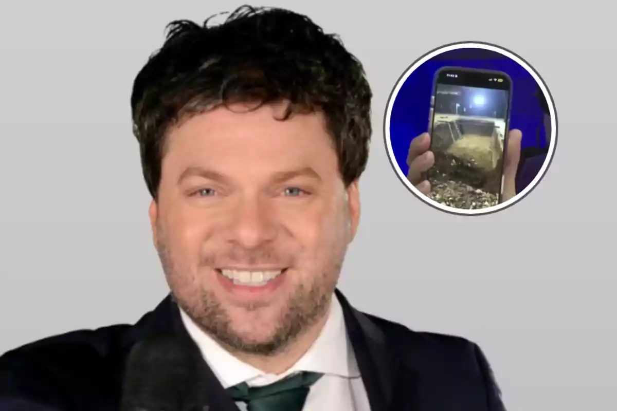 A smiling man with a beard and dark hair, dressed in a suit and tie, holds a microphone; in a circle next to him, a mobile phone is shown with an image of an excavation site.