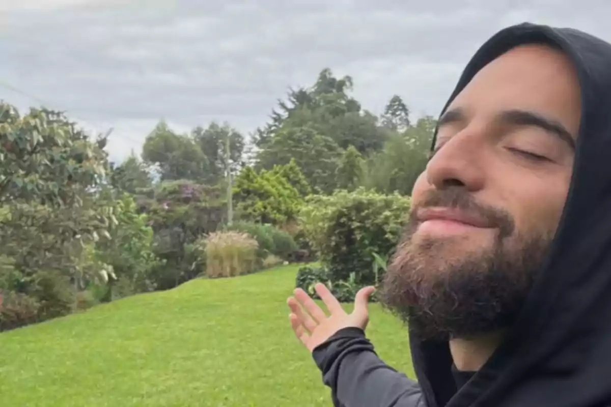 Un hombre con barba y capucha negra disfruta de un momento al aire libre con los ojos cerrados y los brazos extendidos en un entorno verde y natural.