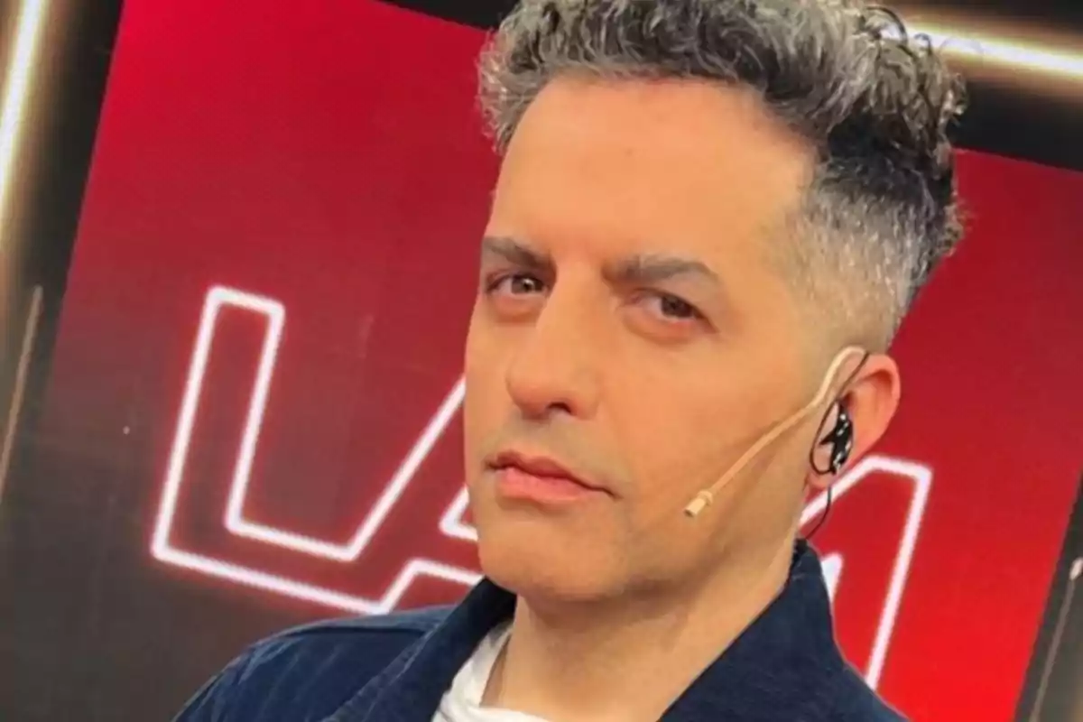 A man with curly gray hair wears a headset microphone in front of a red background with white letters.